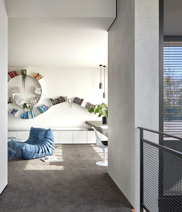 A bright contemporary living room viewed from an adjacent circulation area, featuring a sculptural white built-in media/storage wall with curved open shelving filled with books, low white cabinetry, a plush blue lounge chair, grey carpet, and a small white pedestal side table. A concrete-toned structural wall and black mesh stair or landing balustrade frame the foreground, with strong daylight entering through horizontal blinds.