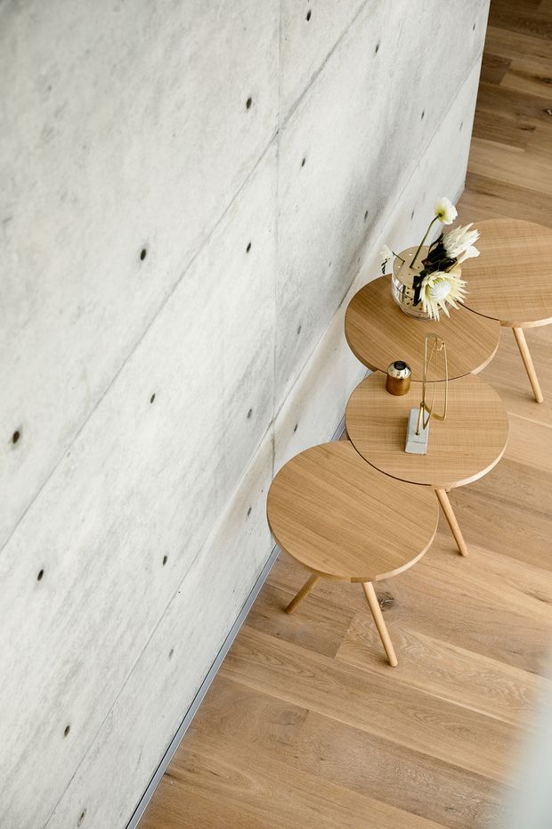A minimalist interior detail showing a row of round light-wood nesting side tables arranged along a pale exposed concrete wall. The space has warm timber flooring, simple tapered timber legs, and sparse decorative objects including a vase of white flowers and a small metallic accessory. The composition emphasizes clean lines, natural wood, concrete texture, and restrained contemporary styling.