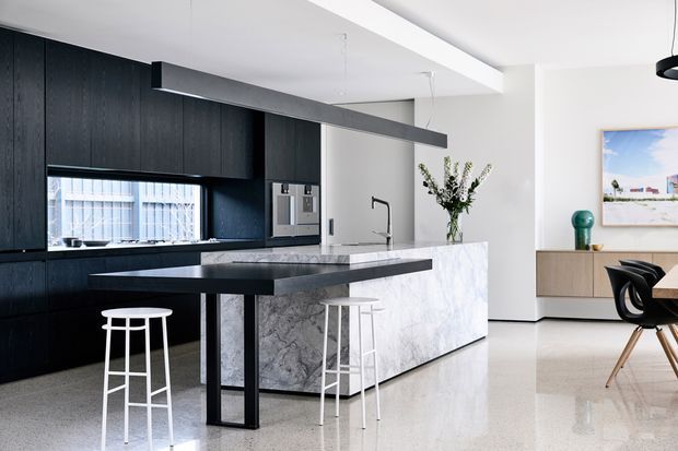 A bright, open-plan contemporary kitchen and dining area with a dramatic black kitchen wall and cabinetry, a large white and grey veined stone island, and a projecting dark island bench with white stools. The space has polished pale flooring, white walls and ceiling, a long slim linear pendant over the island, integrated appliances, and a minimal dining zone with sculptural black chairs and light timber legs. A floral arrangement, artwork and low timber joinery add restrained decorative elements.