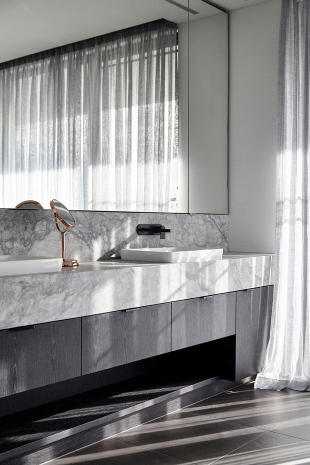 A refined contemporary bathroom vanity with a long grey-veined stone benchtop and integrated backsplash, twin white vessel basins, a wall-mounted matte black tap, and dark timber-look fixed cabinetry below. A large mirrored wall reflects the sheer floor-to-ceiling curtains and bright natural light. Dark large-format floor tiles and a small brushed copper-toned tabletop mirror add contrast.