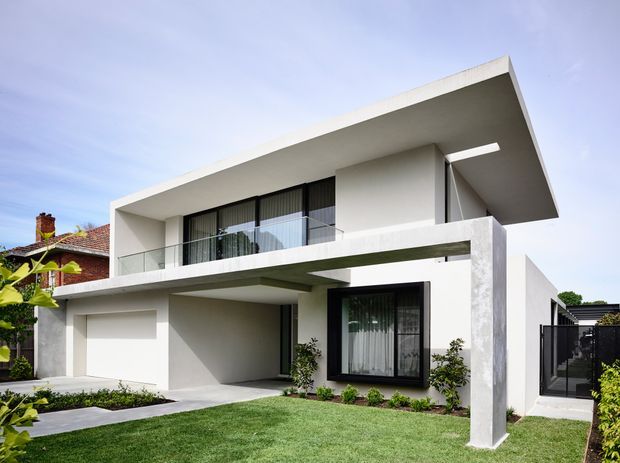 A contemporary two-storey residential house with a crisp white cubic form, deep cantilevered upper roof and balcony, black-framed glazing, and a prominent white structural column. The front garden includes a lawn, paved path, low planting, and a partially enclosed driveway or side access. A neighbouring older brick dwelling is visible at the left edge.