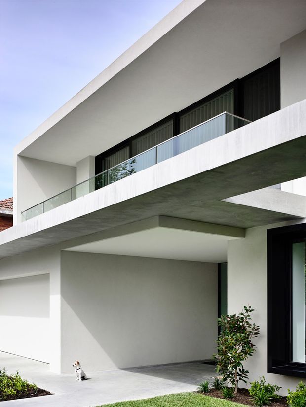 A contemporary multi-level residential facade with crisp white rendered walls, deep cantilevered balcony slabs, black-framed glazing, and transparent glass balustrades. The ground level includes a recessed covered entry or parking area, while a small landscaped garden and driveway sit in front. Strong geometric lines, exposed concrete soffits, and minimal detailing define the exterior.