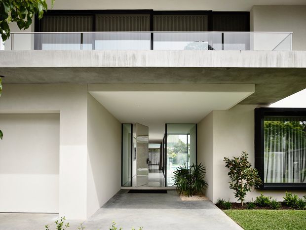 Front exterior view of a contemporary two-storey residence with a broad projecting upper balcony, clear glass balustrade, white rendered walls, dark-framed glazing, and a recessed entry beneath a deep concrete canopy. A simple landscaped garden and paved approach frame the entrance.