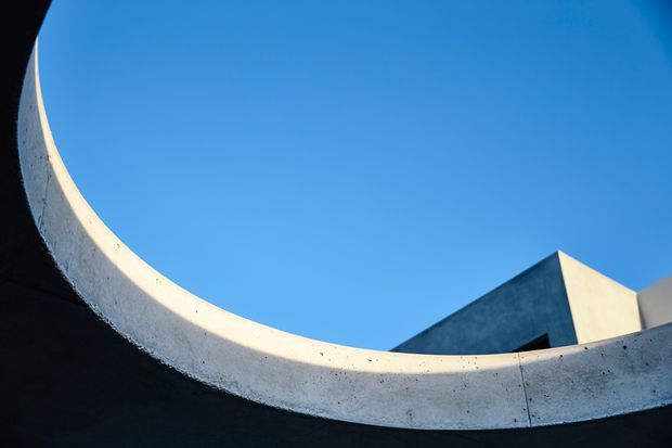Abstract architectural exterior view looking upward beneath a sweeping curved concrete roof or wall edge, with a sharply angled dark and light geometric structure visible against a clear blue sky. The image emphasizes sculptural forms, strong contrast, and minimalist modern construction.