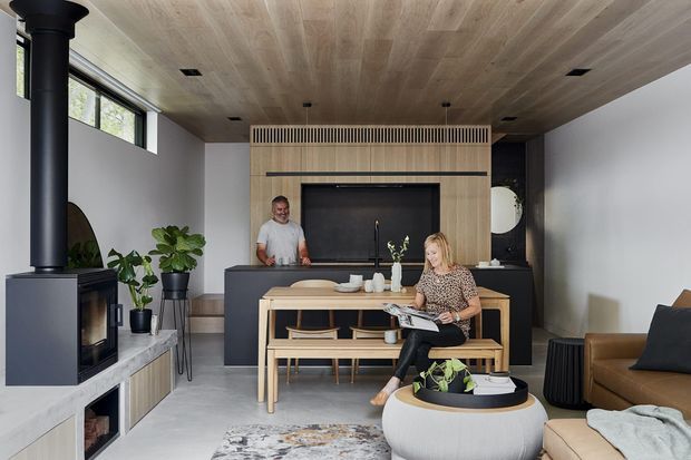 A refined open-plan living and dining space with a dramatic black fireplace and chimney at left, a warm timber-lined ceiling, pale timber feature wall and built-in kitchen cabinetry at the rear. A light timber dining table and bench sit centrally, with a brown sofa, sculptural coffee table, indoor plants and neutral furnishings creating a warm contemporary interior.