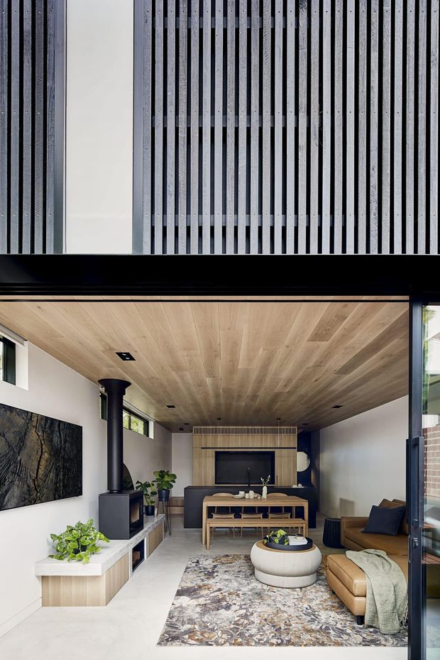 A refined contemporary living room viewed from an open glazed facade. The room features a warm timber-lined ceiling, pale walls and flooring, a dark built-in media wall with vertical slatted detailing, a black cylindrical fireplace flue, low timber joinery, a dining table and chairs, a caramel leather sofa, a round upholstered coffee table, indoor plants and a large patterned rug. Strong vertical architectural screening is visible above the opening, and extensive glazing frames the space.