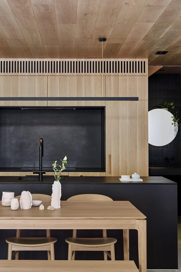 A refined contemporary kitchen and dining area with extensive light timber wall and ceiling panelling, black cabinetry and benchtops, a large dark splashback behind the cooktop, and a timber dining table with matching stools. A linear black ventilation grille and minimalist pendant lighting add strong graphic detailing, while small ceramic and plant accents soften the restrained palette.
