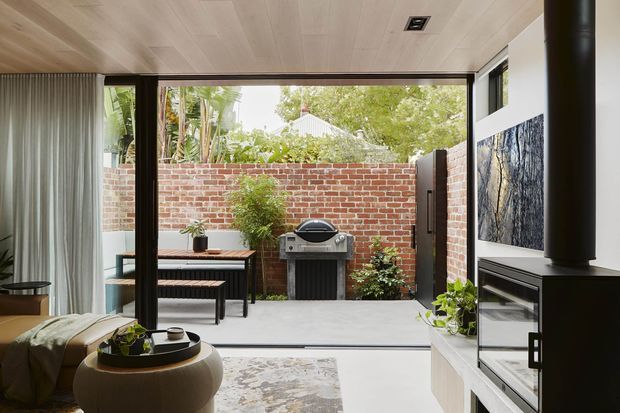 A contemporary living room opens directly onto a compact paved courtyard through wide black-framed glazed doors. The room has a pale timber-lined ceiling, white walls, built-in white cabinetry, a black cylindrical fireplace, neutral furnishings, and a patterned rug. Outside, a red brick boundary wall encloses a small outdoor dining area and built-in barbecue, with lush planting beyond.