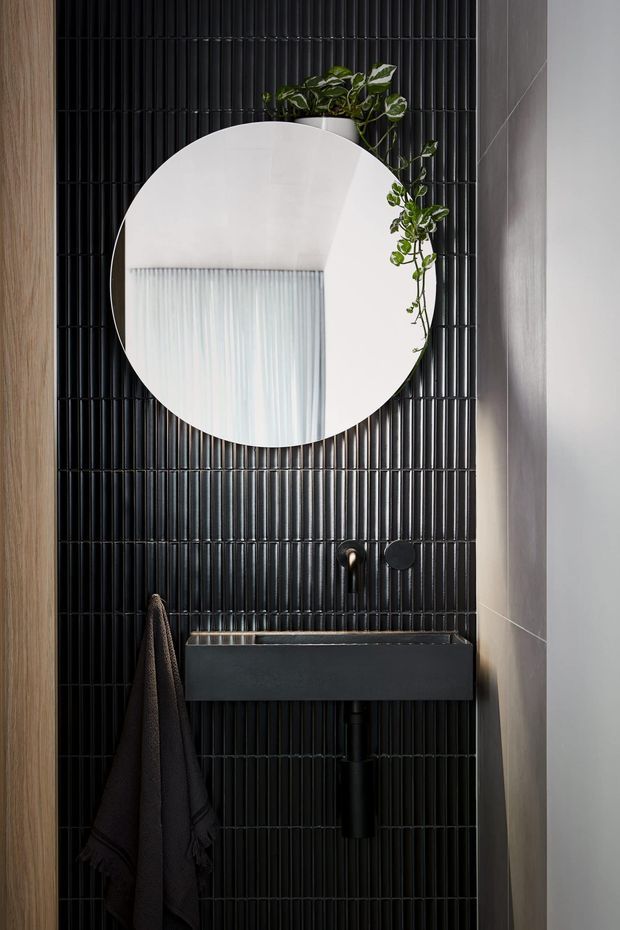 A compact, highly styled bathroom vanity area featuring a wall-mounted matte-black basin with black tapware, a large round frameless mirror, and a trailing green plant. The backdrop is clad in glossy black narrow vertical tiles, framed by warm timber on the left and a smooth light-grey wall on the right. A dark towel hangs beside the vanity, creating a dramatic, refined, contemporary look.