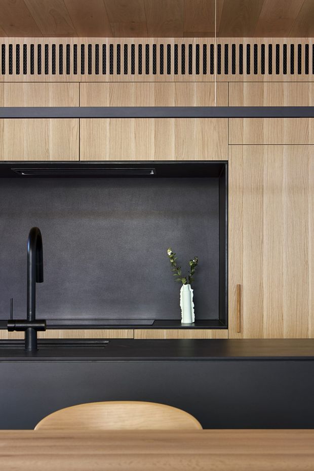 Close-up view of a refined contemporary kitchen featuring warm vertical-grain timber cabinetry, a deep charcoal benchtop and splashback, integrated black tapware, and a dark linear ventilation grille beneath a timber ceiling panel. A small recessed display niche with a vase adds a subtle decorative detail, while the foreground shows the curved back of a timber dining chair and a timber table edge.