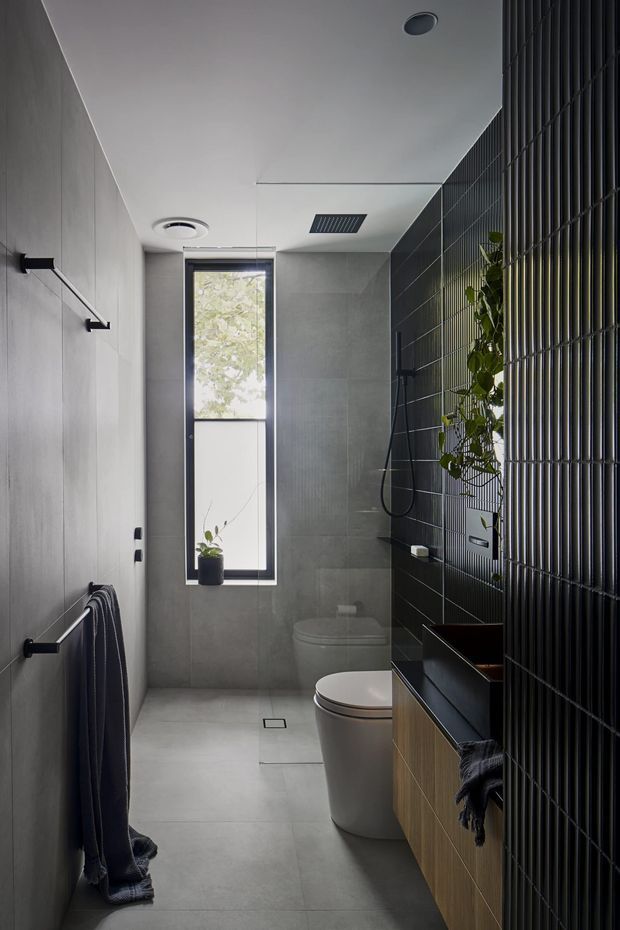 A narrow, contemporary bathroom with large grey wall and floor tiles, a frameless shower area, wall-mounted toilet, freestanding-style toilet, and a timber-fronted vanity with dark benchtop. A black vertical metal screen creates a strong feature wall and partially conceals the vanity, while a tall window provides natural light and greenery is visible inside.