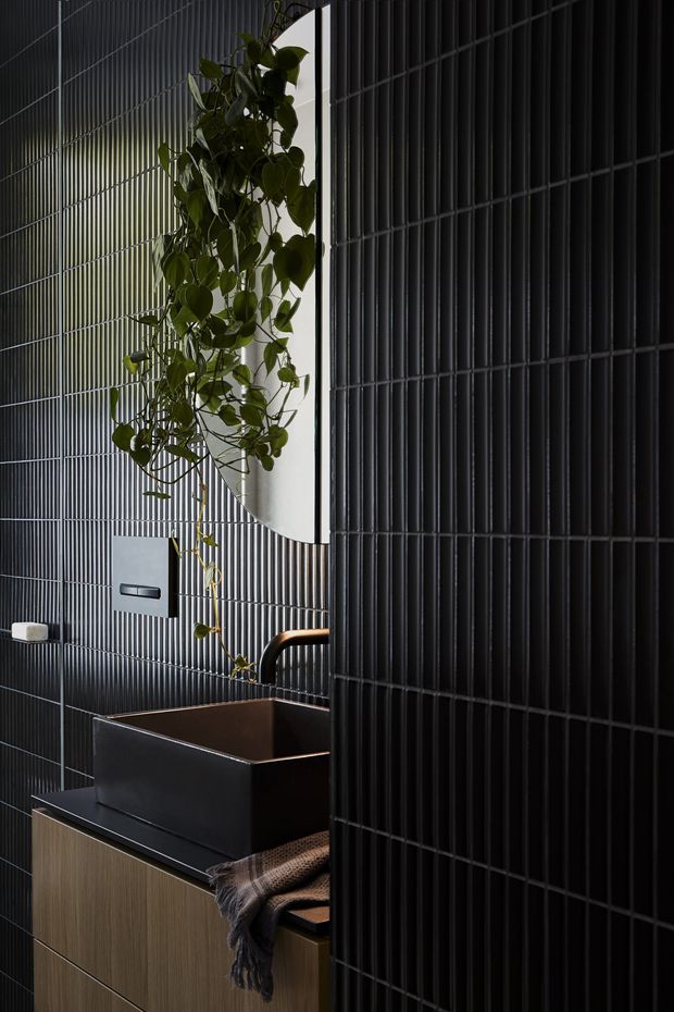 A moody, contemporary bathroom featuring a matte black vessel basin on dark benchtop, vertically ribbed black wall tiles, warm timber-toned vanity cabinetry, and a curved copper-toned tap. A trailing plant hangs beside a partially visible rounded mirror, adding organic contrast to the dark interior.