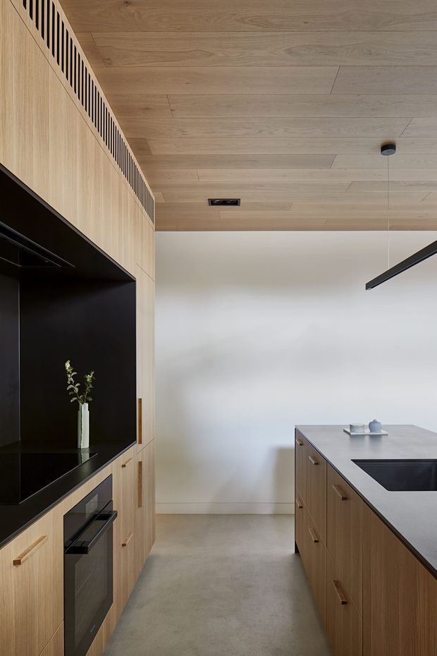A refined contemporary kitchen with full-height light timber cabinetry, integrated black appliances, a large dark-edged island with a pale stone benchtop, and a light timber-panelled ceiling. A black feature wall and linear pendant light create contrast against the otherwise warm, neutral palette.