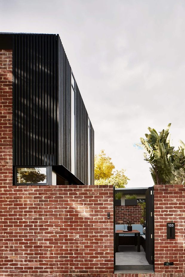 Exterior view of a contemporary urban residence behind a low red brick boundary wall. The facade combines exposed brickwork with a tall black vertical slatted screen and dark-framed glazing. A narrow pedestrian entry opens into a paved courtyard or entry passage, with tropical planting visible behind the wall. The composition highlights the strong contrast between warm brick and black metal detailing.