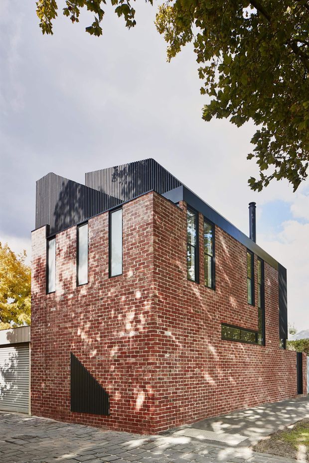 Exterior architectural photograph of a compact multi-storey urban residence. The building features exposed red brick walls, tall narrow windows with dark frames, and a contrasting dark corrugated metal upper volume and roofline. A paved footpath, boundary wall, chimney flue, and mature tree canopy frame the scene.