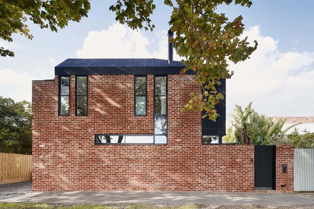 Exterior view of a compact two-storey brick dwelling with a flat-sided rectangular form, dark corrugated metal pitched roof, black-framed narrow windows, and a tall dark entry gate integrated into a brick boundary wall. Timber fencing, corrugated metal fencing, and tropical planting are visible around the property.