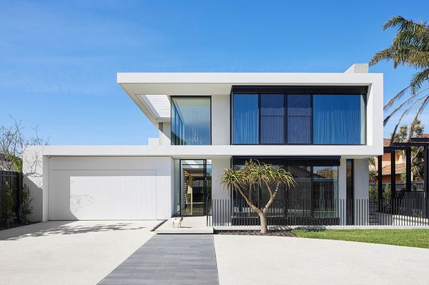 Front exterior view of a contemporary two-storey residence with a crisp white rendered facade, strong rectangular volumes, flat rooflines, extensive dark-framed glazing, a covered upper-level balcony or recess, integrated double garage, front path, lawn, and tropical landscaping. The composition is bright and minimal with a suburban streetscape setting.