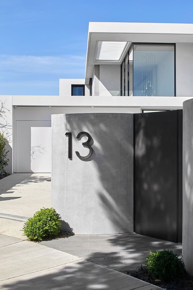 Street-facing view of a minimalist contemporary residence with crisp white rendered forms, broad black-framed glazing, a flat roofline, and a concrete entry wall marked with the number 13. A tall matte-black gate, pale concrete driveway, and sparse clipped landscaping complete the facade. Bright daylight and strong tree shadows create a clean, high-contrast presentation.