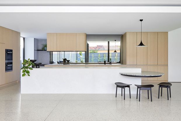 A bright, contemporary open-plan kitchen with pale timber cabinetry, a large white island, integrated appliances, and a narrow raised stone breakfast bar with three dark stools. Black pendant lights, expansive rear glazing, indoor plants, and a polished light terrazzo floor complete the clean, minimalist composition.