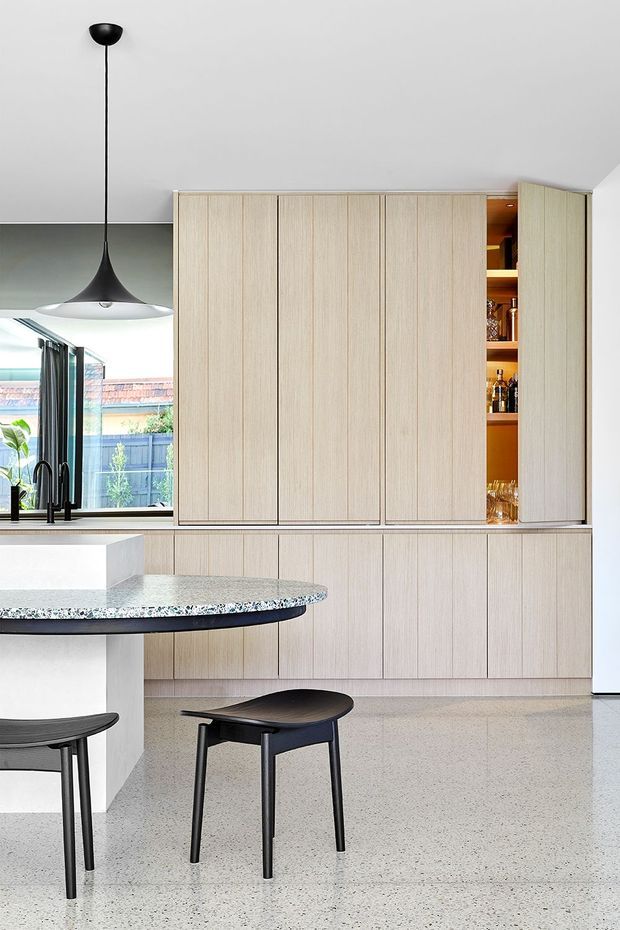 Bright contemporary kitchen with full-height pale timber cabinetry, integrated open bar shelving, a dark stone-speckled island benchtop, black pendant lighting and stools, and polished terrazzo flooring. Large glazing opens the kitchen toward an outdoor area, creating an airy, refined interior.