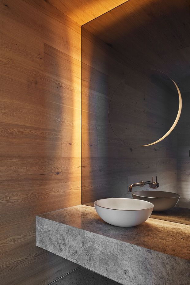 A dramatic, minimalist bathroom vanity featuring dark timber wall panelling, a thick stone countertop and fascia, a white vessel basin with wall-mounted brushed metal tapware, and a large circular mirror. Warm concealed lighting washes the timber and illuminates the vanity, creating a refined, moody atmosphere.