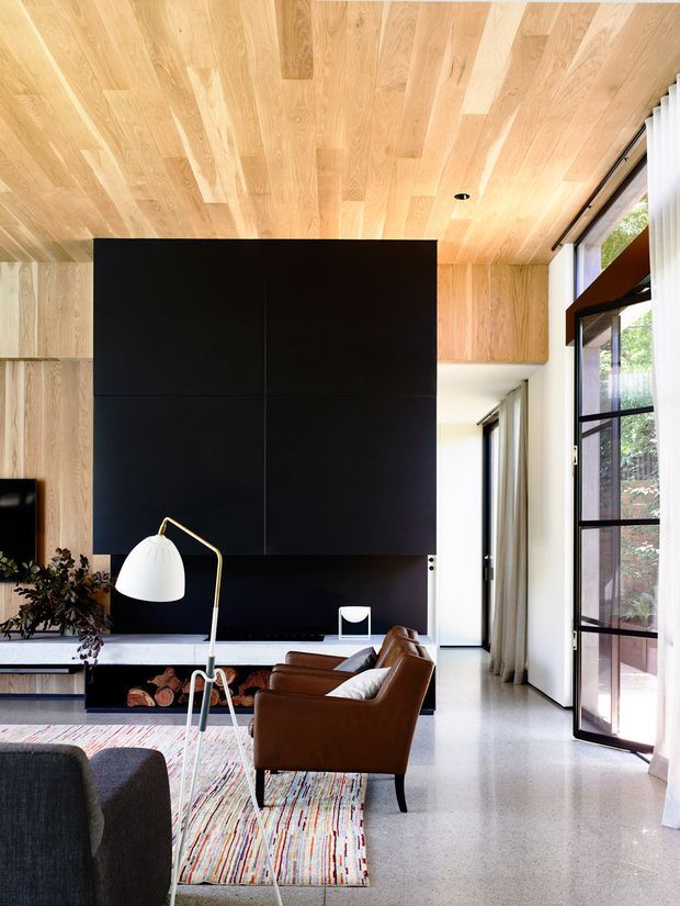 A bright, refined living room with a dramatic black built-in media wall set against extensive light timber wall and ceiling panelling. The room features polished concrete or terrazzo-like flooring, large black-framed glazing with sheer curtains, a brown leather lounge chair, a dark sofa, a sculptural floor lamp, and a colourful linear rug. The high timber-lined ceiling and generous glazing create an airy, contemporary interior with strong contrast between warm wood, black joinery, and pale surfaces.