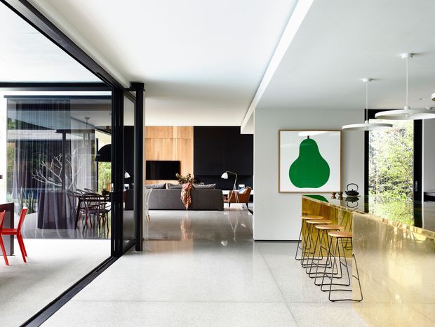 Bright open-plan contemporary interior with a polished light terrazzo floor, white walls and ceiling, and a long pale stone kitchen island with yellow bar stools. Black-framed glass partitions separate the dining area from the living room, which features a grey sofa, dark built-in cabinetry, and timber wall panelling. Large windows bring in views of greenery, while a bold green artwork and sculptural pendant lighting add graphic accents.