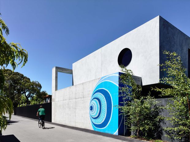 Exterior view of a stark contemporary cubic concrete building with a tall white rendered facade, dark side wall, circular window, and a vivid blue geometric mural on the street-facing wall. The building sits along a paved urban or suburban street with mature landscaping and a cyclist passing by.