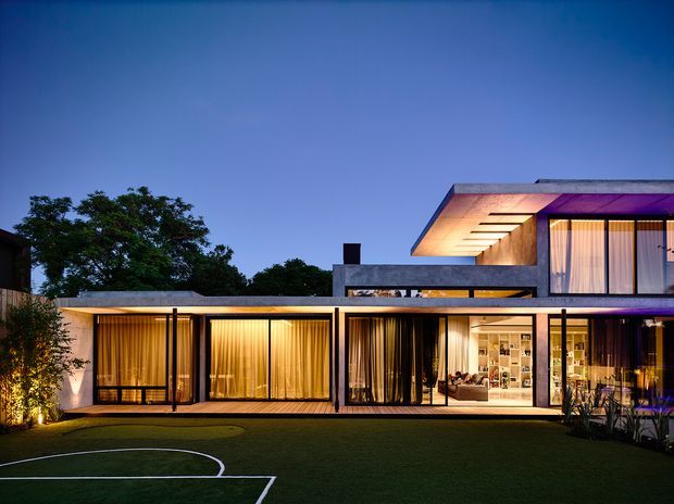 Twilight exterior view of a contemporary two-storey residence with a low horizontal ground-floor wing, projecting upper volume, extensive floor-to-ceiling glazing, dark slender frames, warm interior lighting, and a broad landscaped lawn marked as a sports court.