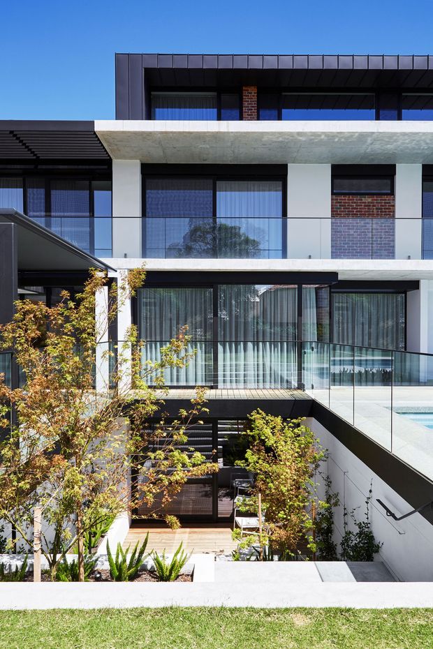A contemporary multi-level residential courtyard facade viewed from outside, featuring crisp white rendered walls, exposed red brick sections, dark metal cladding and roof edges, extensive black-framed glazing, transparent glass balustrades, and a substantial concrete slab. Landscaped planting surrounds a lower-level entry and stair, while a swimming pool is partially visible on the upper terrace. The composition is bright and architectural, with strong horizontal lines and a layered courtyard arrangement.