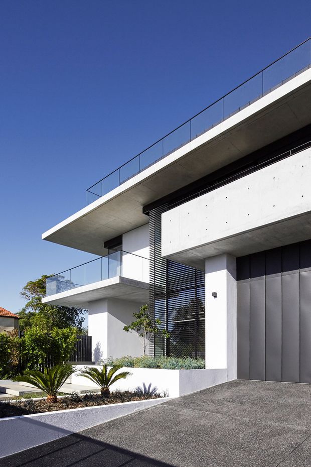 Contemporary multi-level residential facade with crisp white rendered walls, projecting flat concrete roof slabs and balconies, frameless glass balustrades, dark vertical screening, and a large dark garage door. The building is photographed from the driveway, with minimalist landscaping and a clear blue sky.