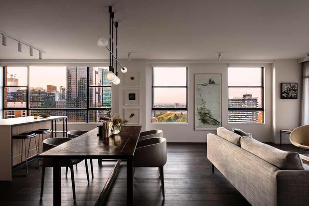Open-plan contemporary apartment interior combining a dining area, kitchen peninsula and living room. The space features large black-framed windows with city views, dark timber flooring, a dark timber dining table, upholstered dining chairs, a neutral sofa, minimalist track lighting and pendant globe lights, with framed artwork and built-in display niches.