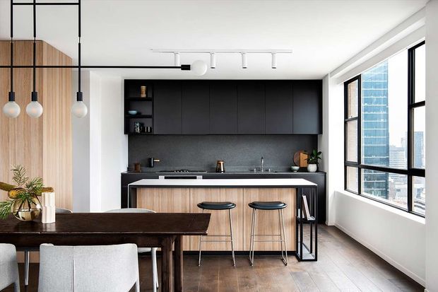 Bright contemporary open-plan kitchen and dining area in an apartment or urban residence. The kitchen features a long dark charcoal island with timber cabinetry, black upper cabinets, a dark textured splashback, integrated appliances, and two black stools. A timber dining table and upholstered chairs occupy the foreground. Large black-framed windows provide abundant daylight and an urban high-rise outlook. Minimal linear track lighting and pendant lights reinforce the clean modern fit-out.