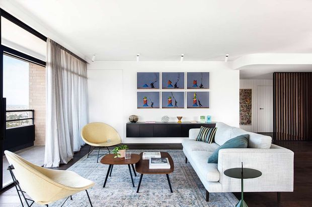 Bright contemporary living room with floor-to-ceiling glazing and sheer curtains, a large pale sectional sofa, sculptural lounge chairs, nesting coffee tables, a long dark built-in sideboard, and a patterned area rug. White walls and ceiling create an airy setting, with colourful framed artwork and a dark vertical timber screen adding visual interest.