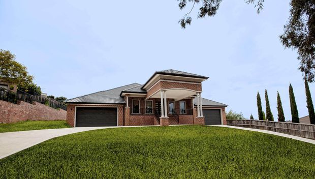 Exterior view of a large red-brick suburban house on a gently sloping lawn. The home has a low-pitched tiled roof, multiple gabled and hipped roof sections, a prominent covered entry porch supported by white columns, dark garage doors, and a raised brick retaining wall with fencing. Tall narrow evergreen trees and mature landscaping frame the property.