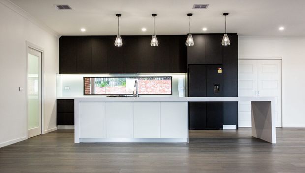 A bright contemporary kitchen with a large white island, dark full-height cabinetry, integrated appliances, a mirrored or glazed splashback, pendant lights, and grey-brown timber-look flooring. The open room also includes white paneled doors and a geometric island support detail.