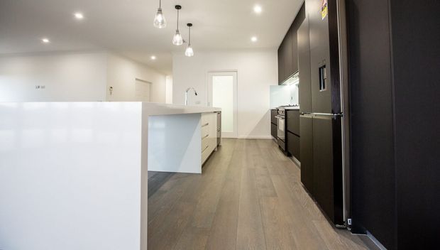 A contemporary open-plan kitchen with a large white island, dark full-height cabinetry and integrated refrigerator along the right wall, stainless-steel appliances, pale wood-look flooring, white walls, recessed ceiling lights and pendant lights over the island.