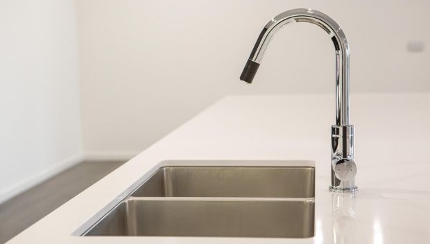 Close-up of a modern kitchen sink with a double stainless-steel basin and a tall curved chrome mixer tap set into a white benchtop. The surrounding space is minimal and softly lit, with a neutral white and grey palette.
