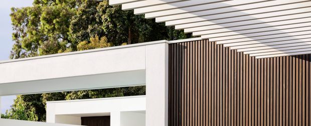 Close-up exterior architectural view of a contemporary residence, featuring crisp white rendered walls, deep rectangular openings, a projecting white pergola with closely spaced horizontal slats, and a tall vertical timber-look screen forming the facade. Mature greenery is visible behind the structure.