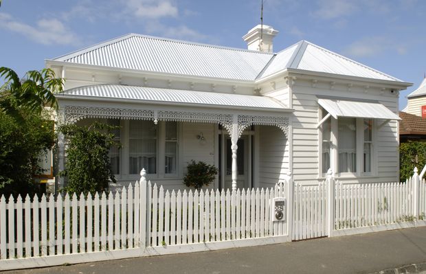 Street-facing view of a restored single-storey white weatherboard house with a corrugated metal hipped roof, ornate Victorian-era verandah detailing, decorative fretwork, turned posts, and a white picket fence. The home sits along a paved urban or suburban street under a bright sky.