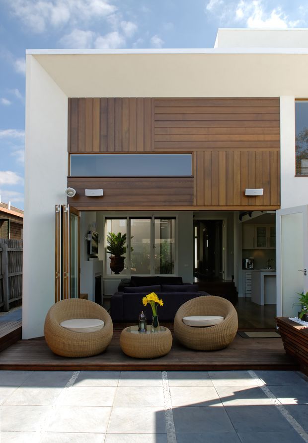 Exterior view of a contemporary two-storey residence opening onto a timber deck and paved patio. The facade combines white rendered walls with extensive warm timber cladding, horizontal and vertical panel arrangements, and large glazed openings. Woven outdoor lounge chairs and a low table sit on the deck, while the open doors reveal a living area and compact kitchen behind.