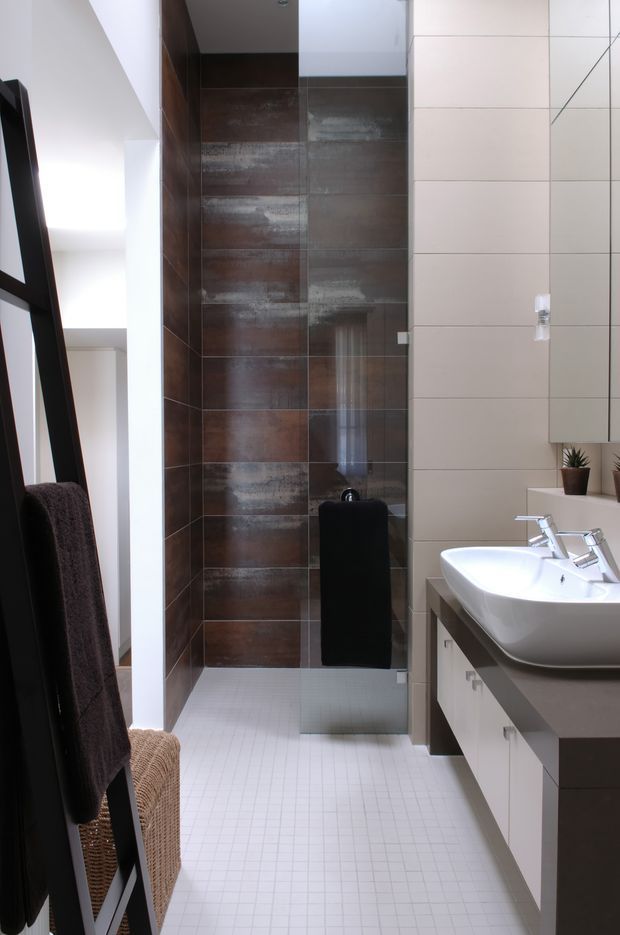 A contemporary bathroom with a long, narrow layout. The space features a walk-in shower lined with dark brown metallic-look rectangular tiles, pale large-format wall tiles, and a white small-grid tiled floor. A wide white double vanity with chrome tapware and light cabinetry occupies the right side, while a dark ladder-style towel rack and woven stool sit on the left. The room is bright, clean, and materially contrasting.