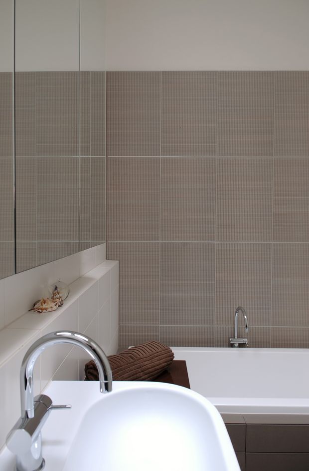 A compact contemporary bathroom featuring a white integrated basin and chrome faucet in the foreground, a white bathtub with chrome tapware, and a full-height wall finished in small taupe-grey mosaic tiles. White tiled ledges and partial mirrored surfaces add brightness, while a dark wood-toned vanity or shelving element provides contrast.
