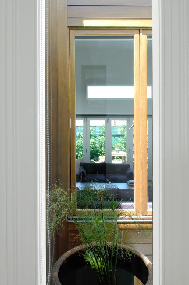 A narrow interior view through a timber-framed glazed opening, looking toward a bright living room with a dark sofa and large windows. The foreground includes a tall indoor planter, while the surrounding opening has white vertical wall or trim panels and warm timber framing. The image emphasises layered glazing, reflected interiors, natural light, and a connection between rooms.