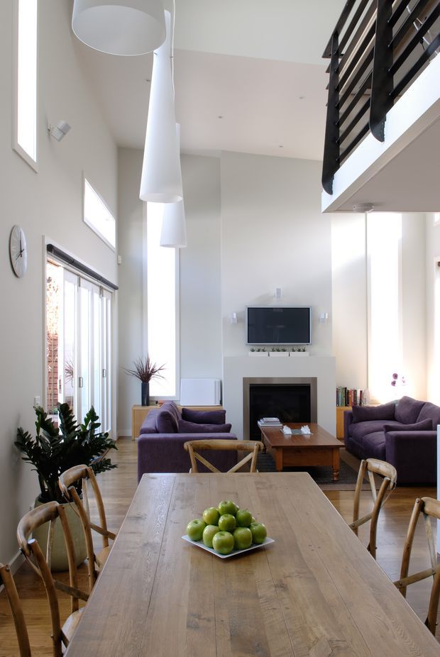 Bright, double-height open-plan living and dining space with a large timber dining table in the foreground, purple sofas, a central fireplace and television, tall glazing, oversized white pendant lights, and a black horizontal-slat mezzanine balustrade. The room has pale walls, timber flooring, abundant natural light, and indoor plants.