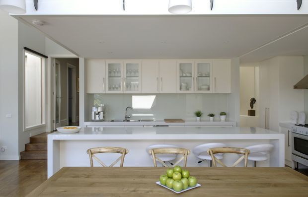 Bright open-plan kitchen and dining area with a large white island and integrated breakfast bar, white shaker-style cabinetry including glass-front display doors, pale benchtops, stainless-steel sink and appliances, and timber flooring. A rustic timber dining table with woven-back chairs occupies the foreground, while steps and a hallway lead away on the left. The space is light, clean, and inviting with a restrained coastal-contemporary feel.