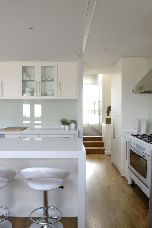 Bright, contemporary white kitchen with extensive built-in cabinetry, glass-front display cupboards, a pale glossy splashback, white island bench with curved bar stools, and a freestanding stainless steel cooker beneath a rangehood. Warm timber flooring leads toward a hallway and short set of timber steps. Recessed ceiling lights and abundant daylight create an airy, clean appearance.
