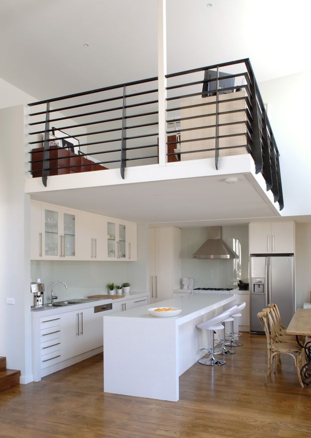 Bright open-plan kitchen with white cabinetry, a large white island with bar seating, stainless-steel appliances and rangehood, and warm timber flooring. A mezzanine-level landing with black horizontal metal balustrading overlooks the kitchen, creating a spacious contemporary interior with high ceilings.