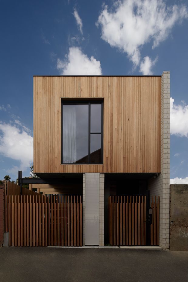 Street-facing exterior of a narrow contemporary multi-storey townhouse. The upper facade is clad in vertical warm timber boards with a large dark-framed window, while the lower level is recessed behind vertical timber slat gates and a pale brick structural wall. The building has a rectilinear flat-roof form and a restrained material palette, photographed against a bright blue sky.
