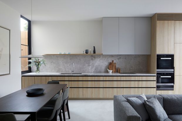 A contemporary open-plan kitchen and dining area with a minimalist, linear composition. The kitchen features light timber-grain cabinetry, pale grey upper cabinets, a large grey stone-look splashback, integrated black appliances, and a long island or peninsula with a slim timber benchtop. A dark dining table and chairs occupy the foreground, while a grey sectional sofa is partially visible at right. Large glazing and timber screening bring natural light into the space.